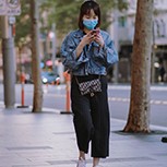Woman in a face mask walks down the street while staring at her phone.