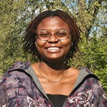 A head and shoulders shot of Nmeso smiling, standing outside in the sunshine in front of green grass and trees.