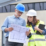 Man and woman on a construction site looking at plans and wearing hard hats. They are in conversation with eachother.