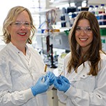 Prof Róisín Owens (CEB) and Dr Amparo Güemes Gonzalez (Engineering) in lab coats, safety goggles and gloves, holding the devices