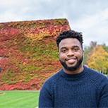 A head and shoulders shot of Dr Ogbeide taken outdoors with red/green foliage visible on the wall of a building behind him