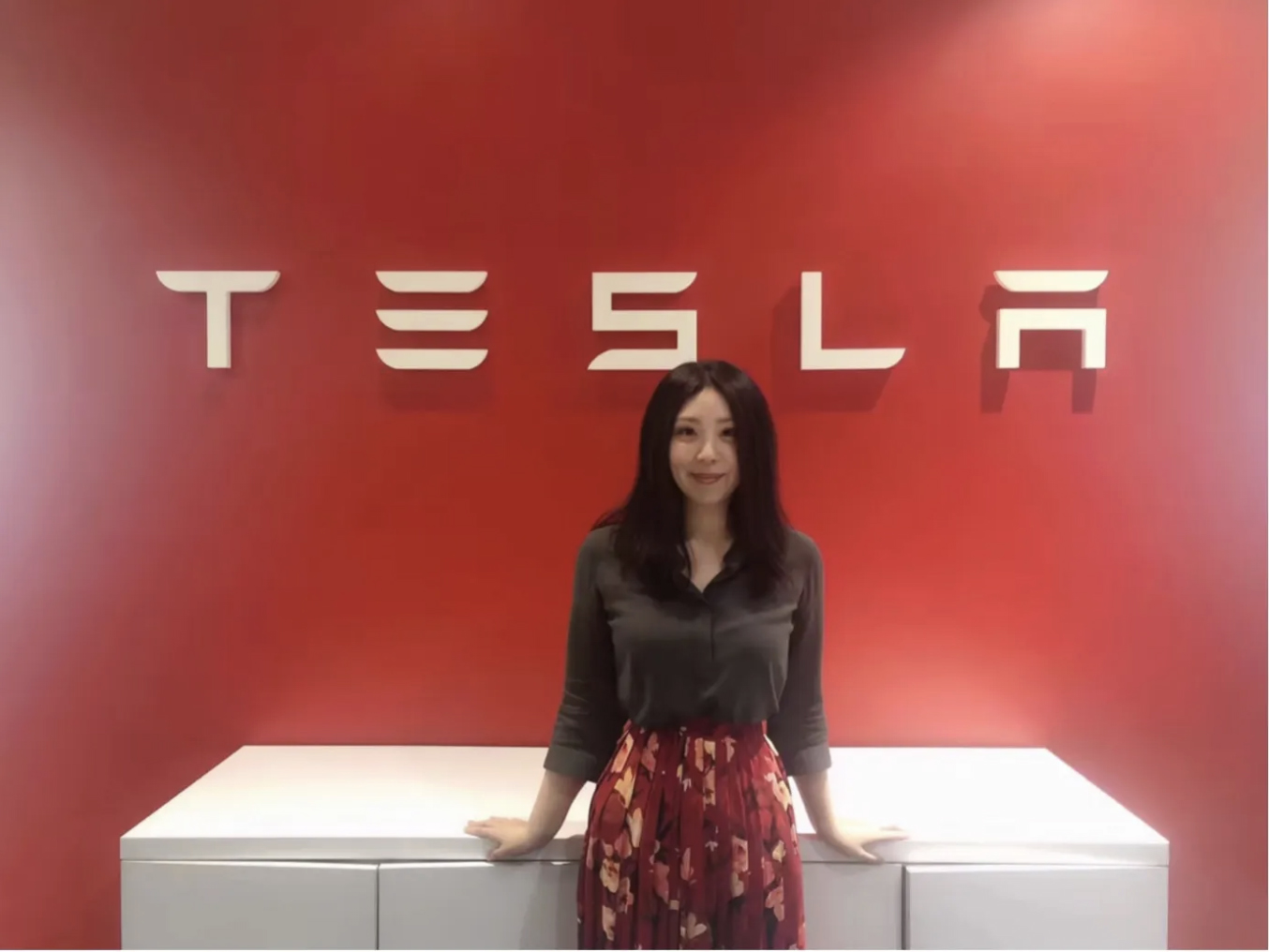 A woman with long dark hair stands smiling in front of a bright red wall featuring the “TESLA” logo in large, silver letters. She is resting her hands on a white counter in front of her.