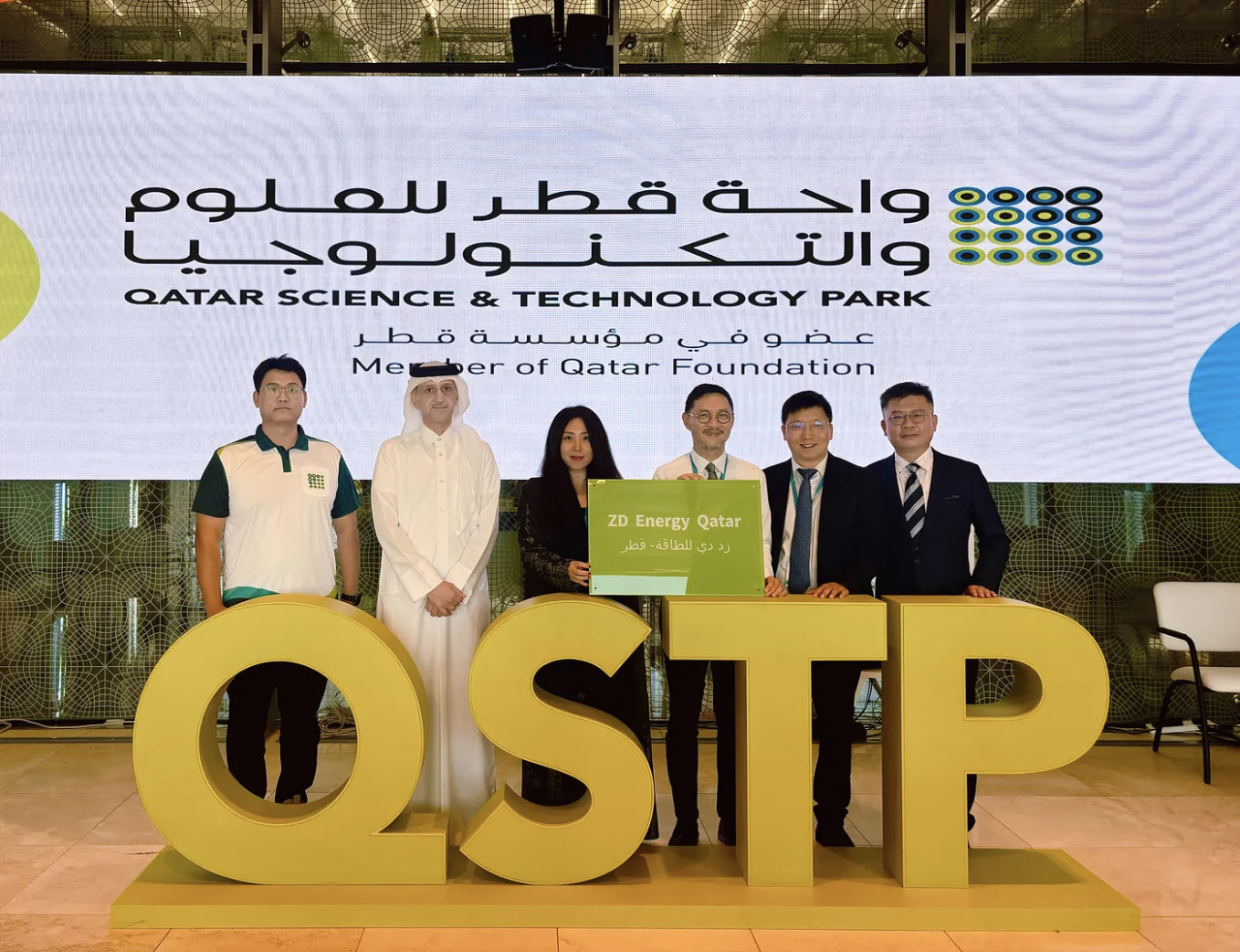 Six people pose for a group photo at a Qatar Science & Technology Park event. They are standing behind large, golden, three-dimensional letters that spell “QSTP”. Five of them are holding a green plaque that reads “2D Energy Qatar”