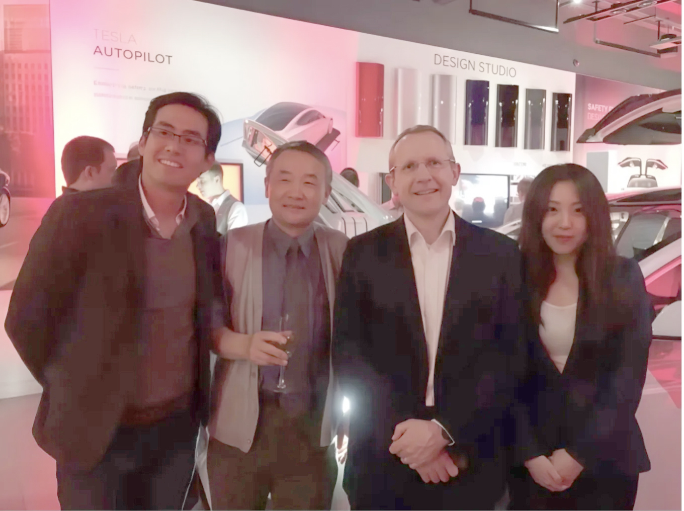 A group of four people — three men and a woman — posing for a photo inside a Tesla showroom. In the background, a white Tesla vehicle is on display with its “falcon-wing” doors open, and wall displays for “DESIGN STUDIO” and “TESLA AUTOPILOT” are visible.