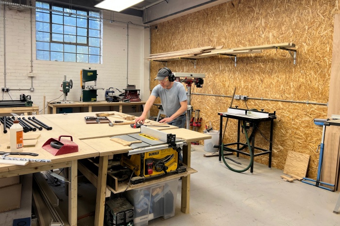 A man in a brightly lit woodworking workshop, wearing safety glasses and ear defenders, leans over a large workbench to work on a wooden project. The workshop is filled with tools and equipment, including a table saw, a bandsaw, and wood stored on wall-mounted shelves against an OSB wall.