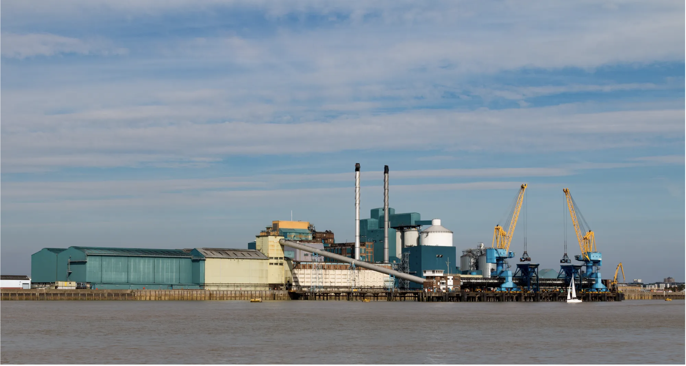 A large, blue and white industrial factory complex on the edge of a wide river under a partly cloudy sky. Several large yellow cranes are visible on the dock.