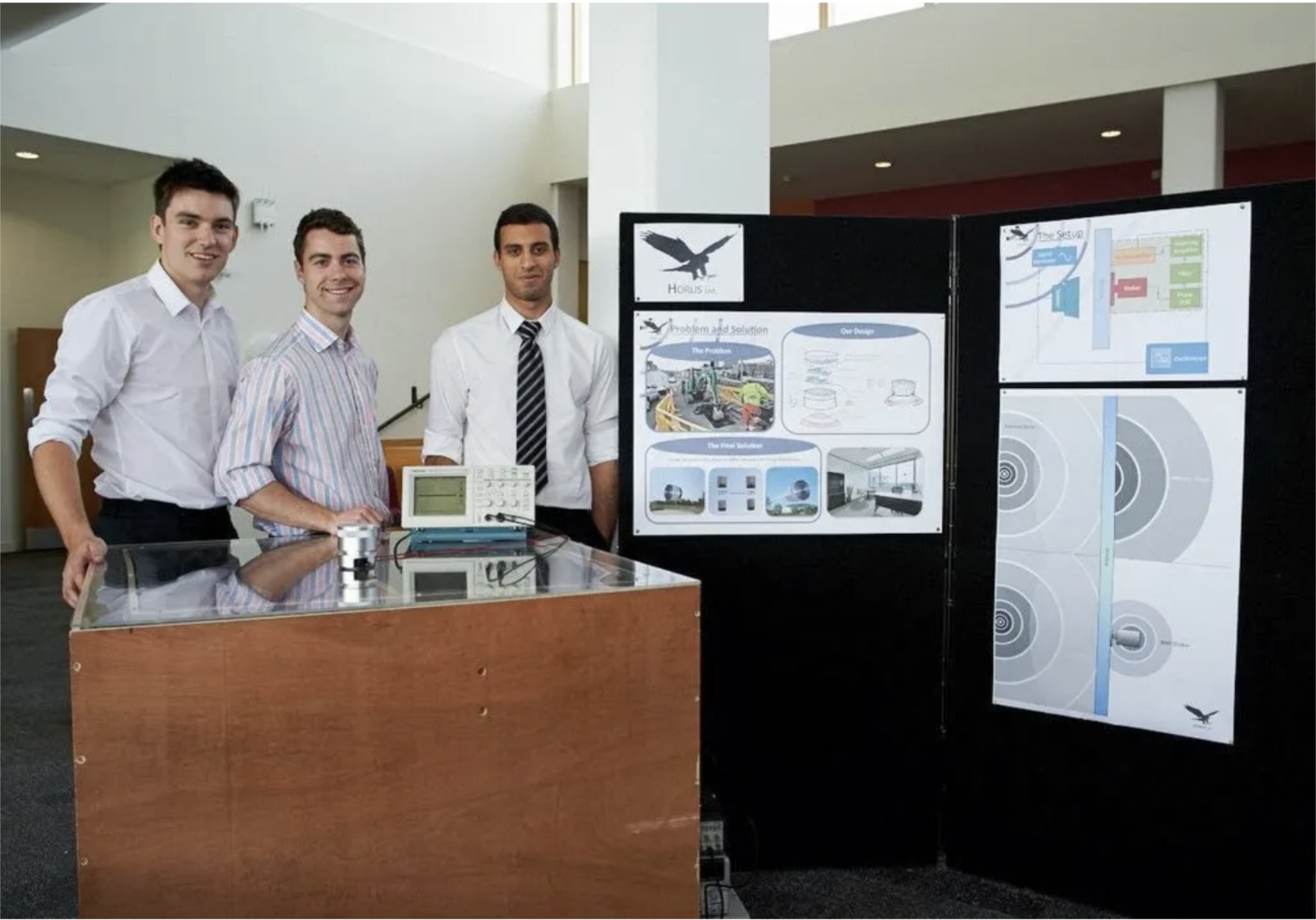 Three young men in smart-casual shirts stand smiling behind a wooden lectern and a black display board featuring project diagrams and posters.