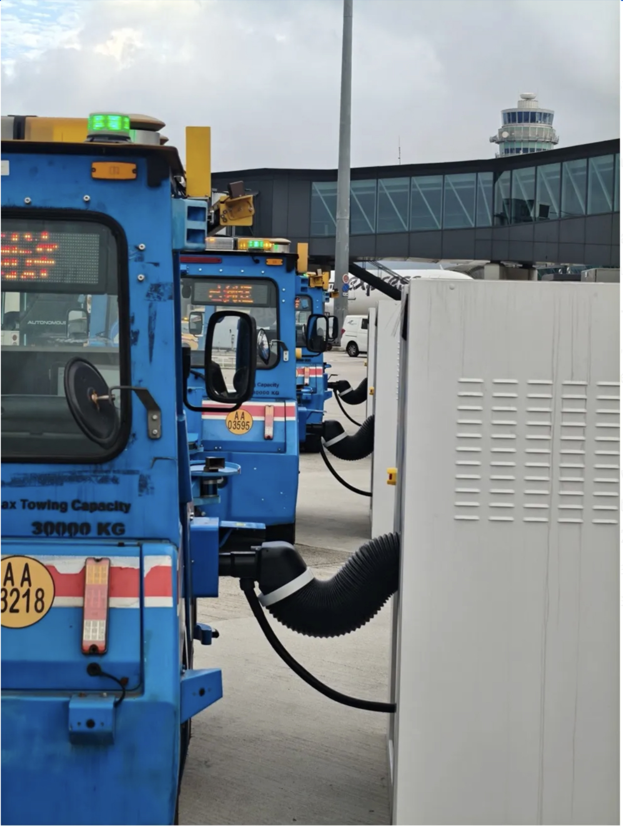 A row of blue electric airport tow tractors parked on the tarmac and connected to white charging stations with black hoses. In the background, an airport terminal and an air traffic control tower are visible under a cloudy sky.